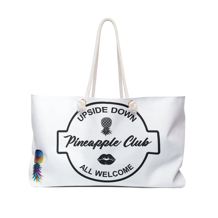 A white weekender bag with a text 'Upside Down Pineapple Club All Welcome' and a colorful pineapple illustration on it, featuring large handles.