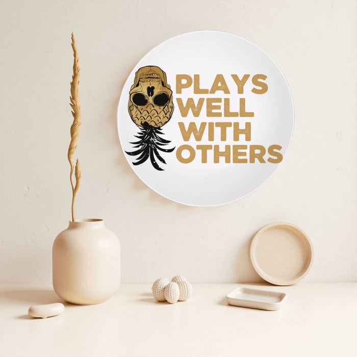 Plays Well With Others decorative plate hanging on a beige wall next to a vase and other house hold good idems.
