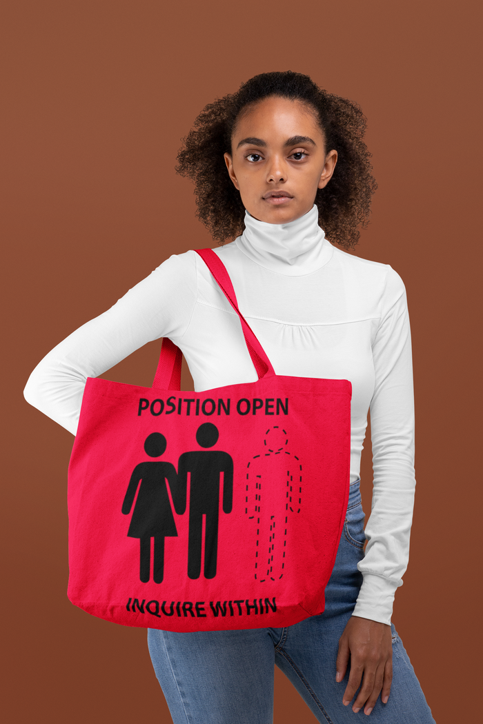 A woman holding a red tote bag with a playful design featuring two cartoon figures and the text 'POSITION OPEN INQUIRE WITHIN'. The bag is designed with a T-bottom shape and has a cream interior lining.